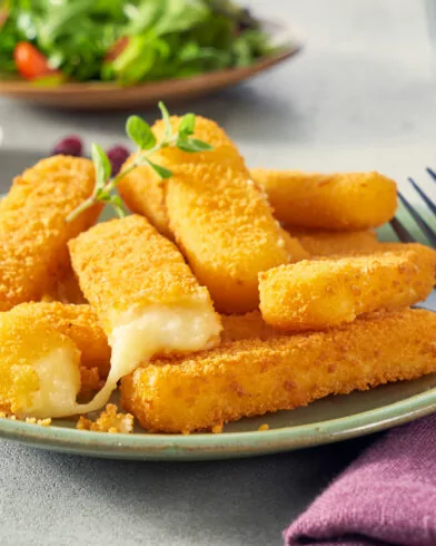 Talíř zlatavých, křupavých tyčinek FRoSTA mozzarella - jedna se sýrem - vedle vidličky, fialového ubrousku, salátu a červené omáčky.