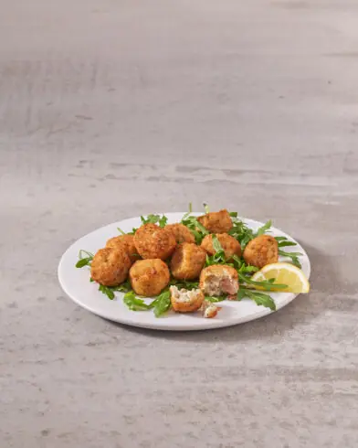 Bílý talíř s několika smaženými kuličkami arancini značky FRoSTA na lístcích zeleně, ozdobený citronem. Jedna z nich je rozkrojená a odhaluje krémovou náplň.
