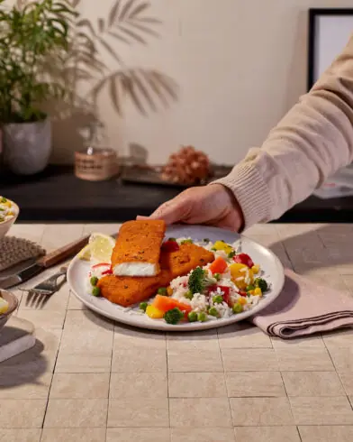 Člověk si servíruje talíř s rybími filé v obalce značky FRoSTA, rýží a zeleninou. Jsou vidět salát, plátky citronu, voda, karafa, rostliny a knihy.